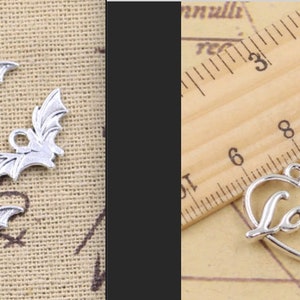 Op de afbeelding: Zilverkleurige metalen bedels. Vier vleermuisvormige bedels met uitgespreide vleugels en een kleine lus om op te hangen. Twee hartvormige bedels met het woord "Love" in cursief schrift. De bedels liggen op een lichtbruin oppervlak.