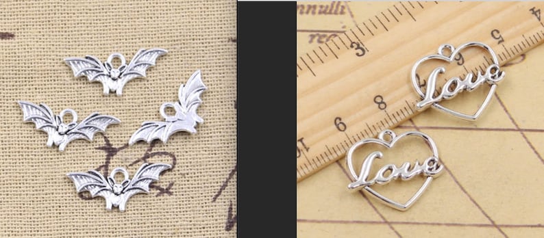 Op de afbeelding: Zilverkleurige metalen bedels. Vier vleermuisvormige bedels met uitgespreide vleugels en een kleine lus om op te hangen. Twee hartvormige bedels met het woord "Love" in cursief schrift. De bedels liggen op een lichtbruin oppervlak.