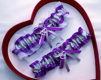 May include: Two purple and silver garters with a silver double heart charm. The garters are displayed in a red heart-shaped box.