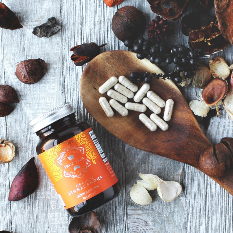 May include: A brown glass bottle with an orange label that reads "True Veda Organic Ashwagandha Dietary Supplement 60 Capsules". The bottle is sitting on a wooden surface with a wooden spoon filled with white capsules.