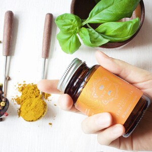 May include: A brown glass bottle with a silver lid containing 60 vegetarian capsules of organic Ashwagandha. The bottle has a label with the text "True Veda" and a floral design.