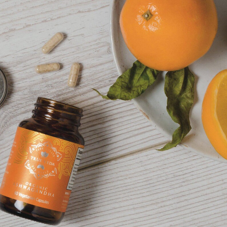 May include: A brown glass bottle with a gold label that reads "Organic Ashwagandha 60 Vegetarian Capsules" and a gold mandala design. The bottle is sitting on a white wooden surface with three capsules and two oranges in the background.
