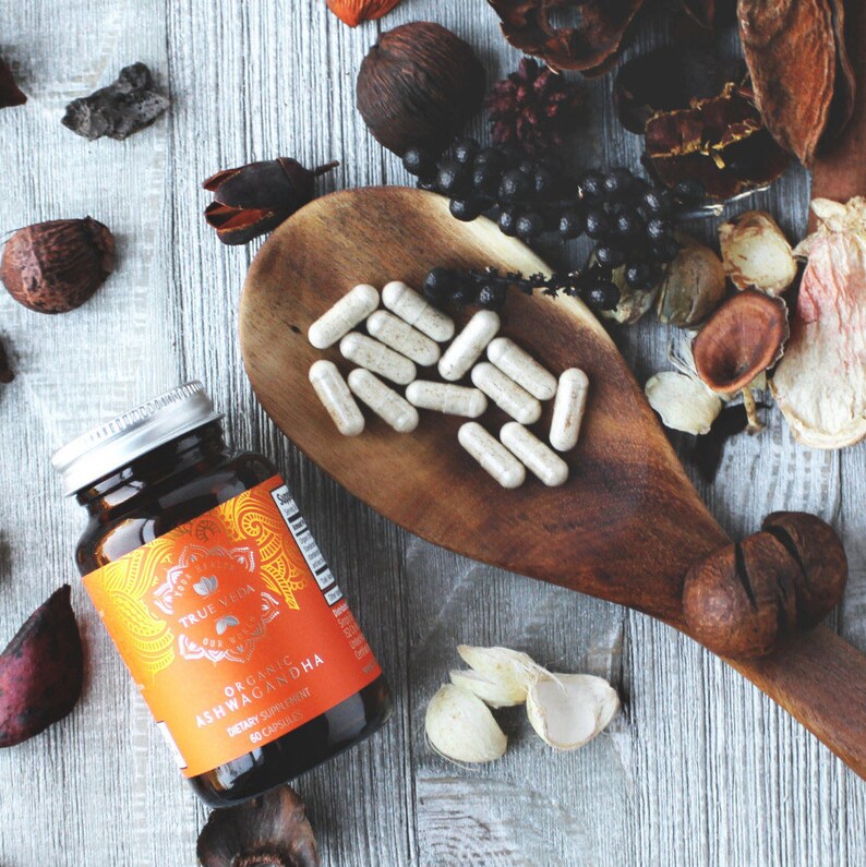 K&ouml;nnte beinhalten: Eine Flasche mit True Veda Organic Ashwagandha Nahrungserg&auml;nzungsmittelkapseln. Die Flasche ist braun mit einem orangefarbenen Etikett und dem Text "True Veda Organic Ashwagandha Dietary Supplement 60 Capsules". Die Flasche steht auf einer Holzoberfl&auml;che mit einem L&ouml;ffel und verschiedenen Kr&auml;utern und Gew&uuml;rzen.