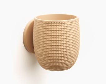 Puede incluir: Una taza de pared de color beige claro con un patrón de cuadrícula texturizado en su superficie exterior. El recipiente tiene una forma redondeada tipo barril y está unido a una base plana circular que se conecta directamente a la pared.