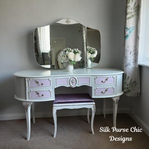 May include: A white vanity table with four light pink drawers, gold-toned hardware, and a tri-fold mirror. A matching stool with a purple velvet seat is tucked underneath. The vanity features ornate gold detailing on the legs and drawer edges.