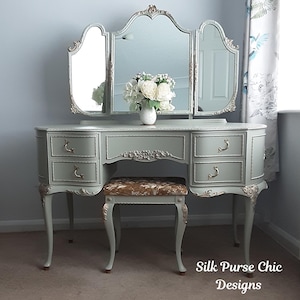 May include: A sage green vanity table with a tri-fold mirror, four drawers, and gold-toned hardware. The vanity features ornate carved detailing and is paired with a matching stool upholstered in a brown, textured fabric. A small vase of white flowers sits on top.