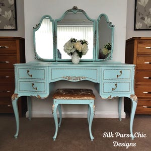 May include: A light blue vanity table with a matching stool and a tri-fold mirror. The furniture features gold-toned decorative carvings on the legs and drawer edges. A white pitcher with flowers sits on the table. Text reads Silk Purse Chic Designs.