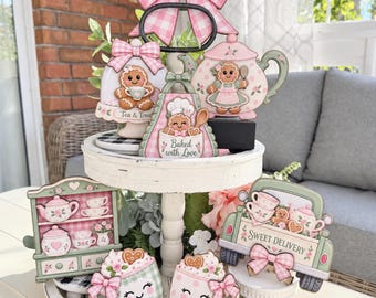 May include: A two-tiered white wooden tray decorated with gingerbread-themed cutouts. Items include a large pink gingham bow, a teapot, a truck labeled Sweet Delivery, mugs with faces, and a hutch. Text reads Tea & Treats and Baked with Love.