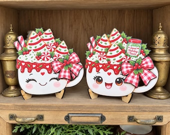 May include: Two decorative wooden mug cutouts featuring smiling faces, red and white checkered bows, and festive toppings including peppermint candies, green holly, and striped straws. One mug displays a jar labeled Sweet Holiday Treats.
