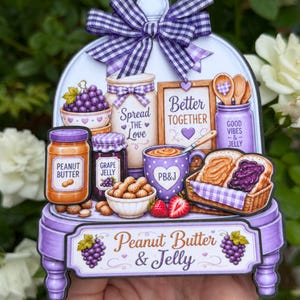 May include: A decorative sign featuring a purple and white gingham bow and a display of peanut butter and jelly themed items. The sign includes jars of peanut butter and grape jelly, a mug, and a basket of sandwiches. Text includes "Spread the Love" and "Better Together."