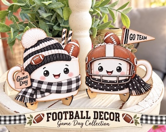 May include: Two wooden cutout decorations shaped like mugs with faces. One mug wears a black and white striped beanie and scarf with a Game Day tag. The other wears a brown football helmet and a black polka dot scarf with a Go Team pennant.