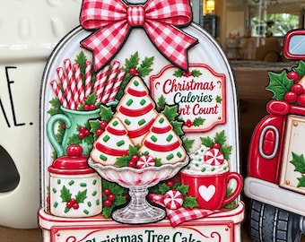 May include: A decorative sign featuring a red and white gingham bow, a glass pedestal dish with three Christmas tree cakes, a red mug with whipped cream, a candy jar, and candy canes. The text reads Christmas Calories Don't Count and Christmas Tree Cake Café.