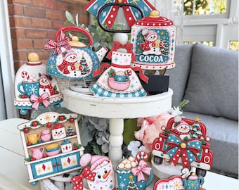 May include: A two-tiered white wooden tray decorated with various Christmas-themed wooden cutouts, including a snowman teapot, a cocoa jar, a kitchen hutch, a red truck, and mugs, all featuring colorful festive patterns and bows.