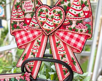 May include: A decorative bow-shaped sign featuring a red and white gingham pattern with illustrations of Christmas tree-shaped cookies, holly berries, and a sign that reads Sweet Holiday Treats, centered with a heart-shaped accent piece.