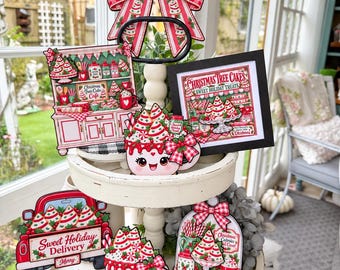 May include: A two-tiered white wooden tray decorated with Christmas-themed signs featuring red and white patterns, illustrations of holiday treats, a red truck with the text Sweet Holiday Delivery, and mugs with faces and Christmas tree cakes.