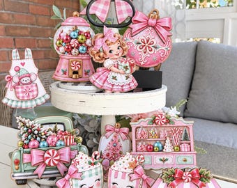May include: A two-tiered white wooden tray decorated with various Christmas-themed wooden cutouts, including a gumball machine, a girl holding a gift, a truck with ornaments, mugs with faces, a candy cart, and a pink bow topper.