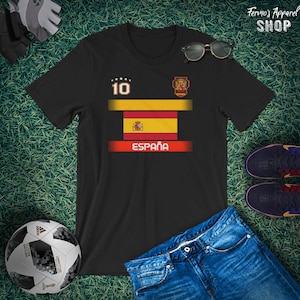 May include: Black t-shirt featuring the Spanish flag design with the word "Espa&ntilde;a" in white. The shirt has the number 10 on the upper left and a crest on the upper right. Sunglasses, a soccer ball, and jeans are also in the image.