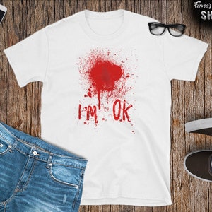 Puede incluir: Camiseta blanca con un diseño de salpicaduras de sangre roja y el texto "I'm OK" en rojo.