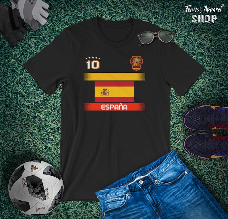 May include: Black t-shirt featuring the Spanish flag design with the word "Espa&ntilde;a" in white. The shirt has the number 10 on the upper left and a crest on the upper right. Sunglasses, a soccer ball, and jeans are also in the image.