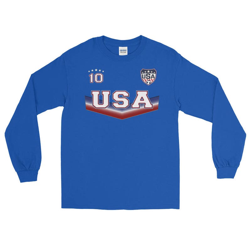 May include: A royal blue long-sleeve t-shirt with a white, red, and blue USA graphic. The graphic includes the word "USA" in white with red and blue accents, a star pattern, and a small USA logo with a soccer ball.