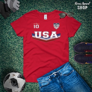 May include: Red t-shirt with a white "USA" graphic and a blue and red chevron design. The shirt also has a white "10" on the chest and a small "USA" logo on the right side.