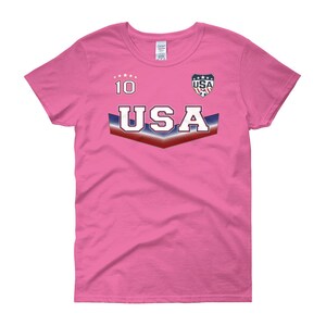 May include: Pink women's t-shirt with a USA soccer jersey design. The shirt has a blue, white, and red chevron with the word "USA" in white letters. The number "10" is printed above the chevron. A small USA soccer logo is printed above the chevron on the right side.