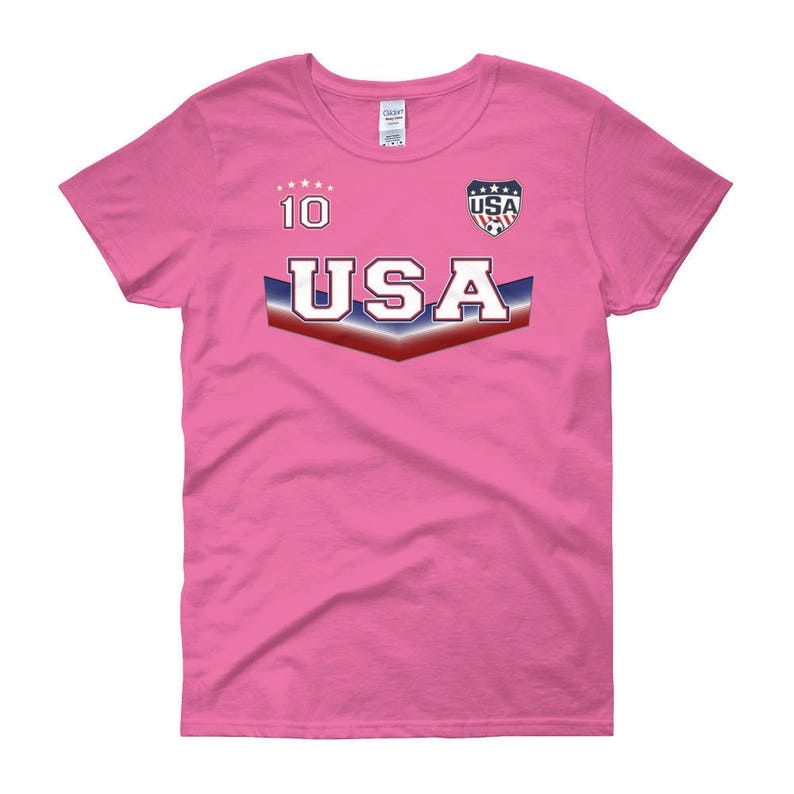 May include: Pink women's t-shirt with a USA soccer jersey design. The shirt has a blue, white, and red chevron with the word "USA" in white letters. The number "10" is printed above the chevron. A small USA soccer logo is printed above the chevron on the right side.