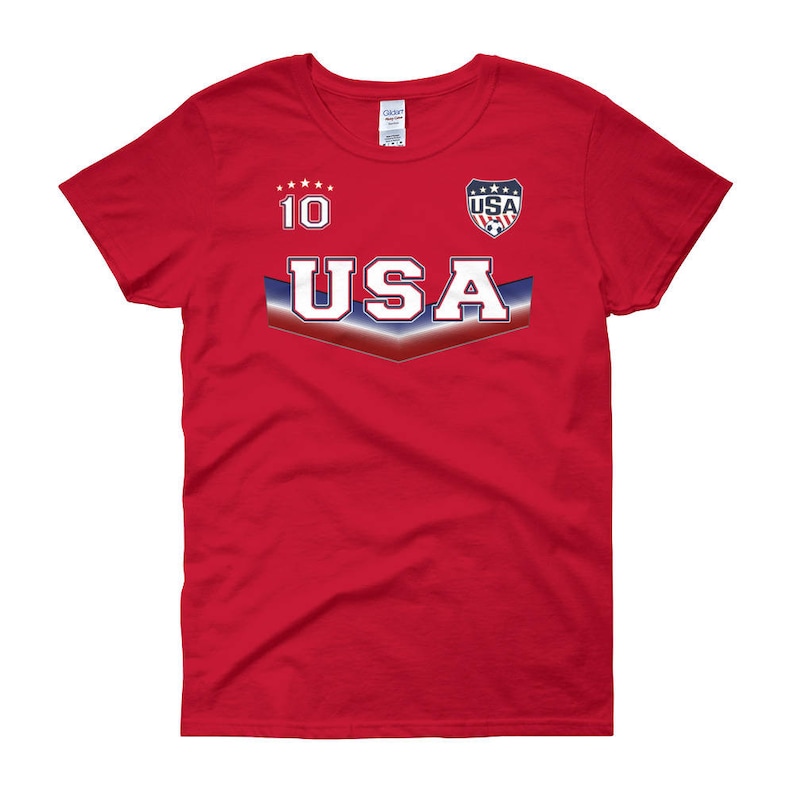 May include: Red women's t-shirt with a white and blue USA soccer jersey design. The shirt has the number 10 and the word USA on the front.