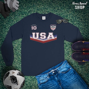 May include: Navy blue long-sleeve shirt with a white "USA" graphic and red, white, and blue stripes. The shirt has a white "10" on the left chest and a small shield with "USA" inside on the right chest.