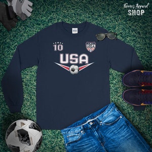 May include: Navy blue long-sleeve shirt with a soccer ball graphic and the text "USA" and "10". The shirt also features a red, white, and blue shield with the text "USA" inside.