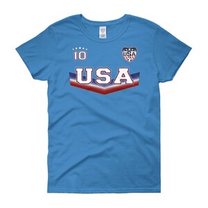 May include: A light blue women's t-shirt with a red, white, and blue USA graphic. The graphic includes the word "USA" in a bold font and a star-shaped design. The number "10" is printed above the graphic.