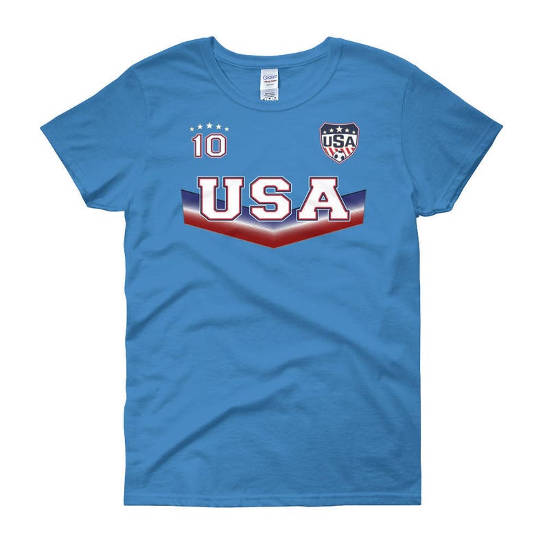 May include: A light blue women's t-shirt with a red, white, and blue USA graphic. The graphic includes the word "USA" in a bold font and a star-shaped design. The number "10" is printed above the graphic.