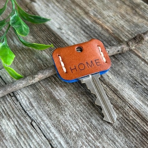May include: A brown leather key cover with the word "HOME" embossed on it. The key cover has a blue border and white stitching. A silver key is inserted into the cover. The key and cover are on a wooden surface.