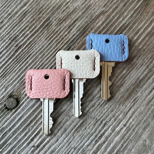 May include: Three keys with leather key fobs in pink, white, and blue. The key fobs are rectangular with a stitched border and a hole for the key ring. The keys are on a weathered wooden surface.