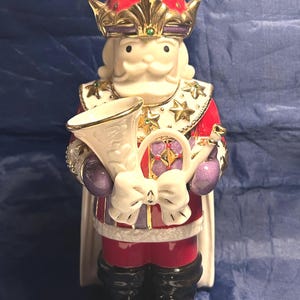May include: A ceramic figurine of a king in a red and white robe, gold crown, and black boots. The king holds a white horn and a purple orb. The figurine has a white beard and mustache, and is decorated with gold accents and stars.