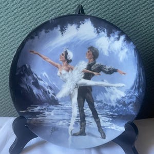 May include: A porcelain plate with a painted image of two ballet dancers in a snowy landscape. The dancers are wearing white tutus and black costumes. The background is a blue and white sky with mountains in the distance.