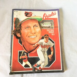 以下が含まれることがあります： 1983年のボルチモア・オリオールズのヴィンテージな野球スコアブック。表紙には、アクション中の野球選手のカラフルなイラストが描かれており、チームロゴと「オリオールズスコアブック '83」の文字が書かれています。