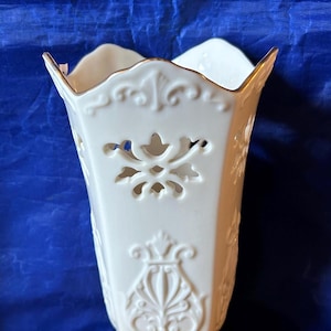 May include: A white ceramic vase with an ornate, embossed floral design. The vase has a flared top with a gold trim and cut-out details. The base is also decorated with embossed patterns. The vase is set against a blue background.