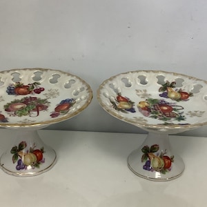 Puede incluir: Dos soportes para tartas de porcelana blanca con ribetes dorados y un diseño de frutas. Los soportes tienen un borde festoneado y un diseño calado.