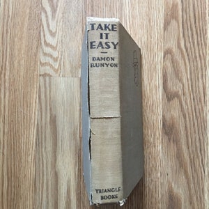 May include: A vintage hardback book titled "Take It Easy" by Damon Runyon. The book cover is beige with black text and a small illustration of a man in a hat. The book is published by Triangle Books.