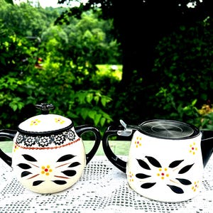 Puede incluir: Dos teteras de cerámica con asas y tapas negras. Una tetera tiene una tapa con un diseño floral negro y un asa negra. La otra tetera tiene una tapa plateada y un pico negro. Ambas teteras tienen una base blanca con diseños florales negros y amarillos.