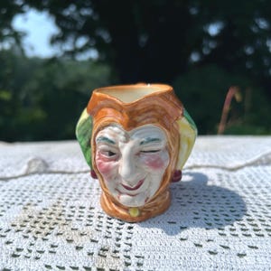 May include: A small, ceramic jug in the shape of a winking jester's head. The jug features a detailed face with rosy cheeks, green and yellow accents on the jester's hat, and a gold-colored collar. The jug is likely a decorative item.