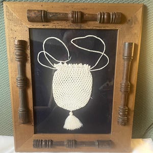 May include: A white crocheted drawstring bag with a tassel hanging from the bottom, framed in a wooden frame with decorative wooden accents.