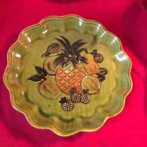 May include: A green ceramic plate with a scalloped edge features a painted design of a pineapple and other fruits. The plate has a glossy finish.