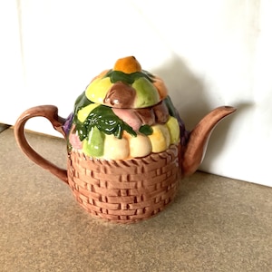 Puede incluir: Una tetera de cerámica con forma de cesta llena de fruta. La tetera está decorada con una variedad de frutas, incluyendo manzanas, peras, uvas y limones. La cesta es marrón y tiene una textura tejida.
