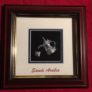 May include: A framed silver-colored metal sculpture of a traditional Arabic coffee pot and cup, set against a black background. The frame is brown with a gold trim and the text "Saudi Arabia" is printed below the sculpture.