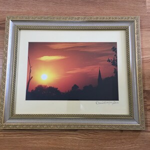 May include: Framed photograph of a sunset over a town with a church steeple in the distance. The sun is a bright orange and the sky is a mix of orange, red, and pink. The signature "David Pemperton" is in the bottom right corner.