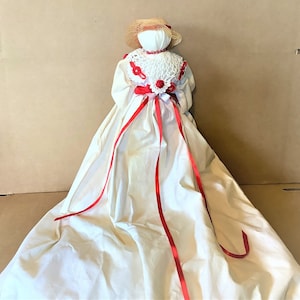 Puede incluir: Una muñeca de tela blanca con una cinta roja y un cuello de crochet blanco. La muñeca lleva un sombrero de paja y tiene una cara de tela blanca.