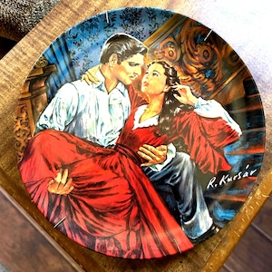 Puede incluir: Plato decorativo con una escena romántica de un hombre que lleva a una mujer por una escalera. La mujer lleva un vestido rojo y el hombre una camisa blanca. El plato tiene un fondo azul y dorado y está firmado "R. Kursár".