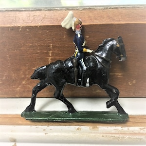 Peut inclure: Un cheval en jouet peint en noir avec un cavalier. Le cavalier porte un uniforme blanc et jaune avec un chapeau blanc. Le cheval est debout sur une base verte.